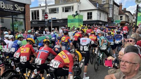 Tens of cyclists sit at the start of the Tour of Britain Men race waiting to get going. Crowds of people watch on from behind railings either side of the road.