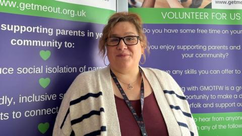 Woman stands in front of some posters - she is wearing a cream and black-striped cardigan and glasses over a burgundy T-shirt. She has large hoop earrings. 