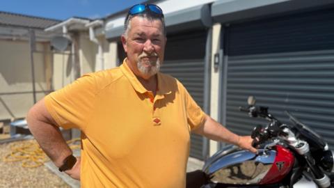 A man with short dark grey hair and a white goatee beard looking at the camera. He is wearing a mustard yellow polo and has sunglasses resting on his head. You can see a red and silver motorbike in the background