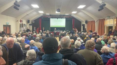 The view from the back of the public meeting looking over rows of seats filled in a community hall - at the front is a stage with a screen and slideshow