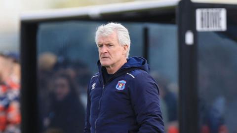 Mark Hughes is wearing a hooded jacket and is standing on the touchline during Carlisle's win at Truro. 
