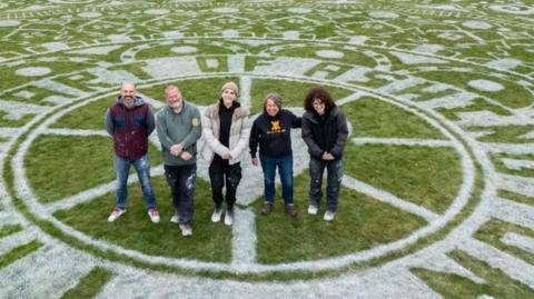 Four smiling people are standing in the central circle of the artwork, made using white marker, that contains the words "Heart of Ashington". It depicts lots of symbols including of ash branches, footballs and miners' wheels. 