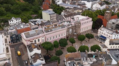 An aerial image of a long building made from pink granite, with white windows. A number of green trees are visible in front of the building.