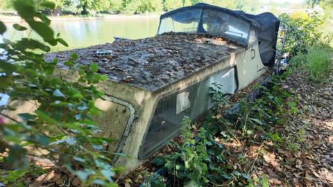 An abandoned boat sits on the edge of the river, covered in leaves and grime. It is half-sunk.