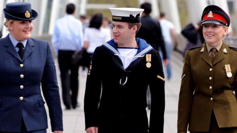 Three uniformed military staff, one man and two women, walk side-by-side across a bridge. One is in a navy blue uniform with a hat and skirt, one is in dark trousers and top with blue collar and white hat, and one is in a brown skirt and buttoned blazer with a hat.