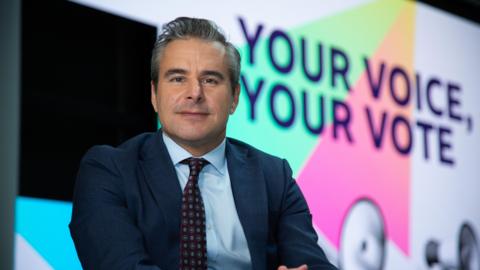 Nick Servini sits in front of Your Voice, Your Vote branding. He wears a suit and tie.