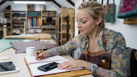 A businesswoman sits in her work premises with an open notebook and her phone displaying a calculator app