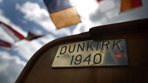 A brass plate reading "DUNKIRK 1940" on the deck of a boat. In the background there are semaphore flags on bunting. 