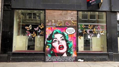 A boarded up shop in Newport with a pop art woman painted onto a board and a speech bubble which reads: "This town has gone to the dogs"