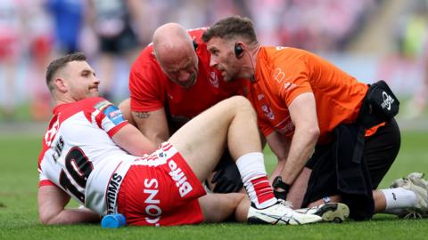 Matty Lees is down on the ground with a rueful look on his face and his right leg bent upwards as two physios treat his left knee which is prone on the ground