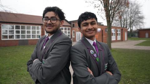 Abdullah and Bakhtiar are standing back to back with their arms folded smiling at the camera. They are wearing their gray school blazers, purple ties and white shirts. 