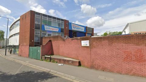 The outside of the Scrapstore building. It is a large brick warehouse with lots of windows on one side and white cladding on the other. It has a faded sign on the front which says 'Children's Scrapstore'. There is a long red brick wall which encloses the car park, directly outside the entrance to the shop.