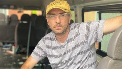 A man wearing a yellow cap and a grey t-shirt sitting in the back of a van.
