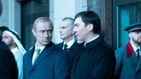 A scene from The Wizard of the Kremlin. Jude Law is dressed as Vladimir Putin, in a suit and with thinning blond hair. He is staring at the man next to him, who is also in a jacket and suit and has dark hair. They are standing outside with various other figures milling about.