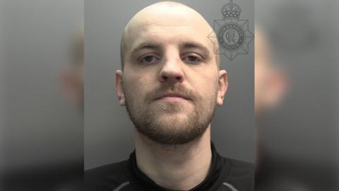 A custody photograph of a man with a shaved head and a brown beard and moustache. He is wearing a black top.