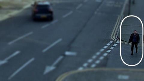 A CCTV image of a man wearing dark clothing walking down a street. The rest of the street is blurred, but the man is circled and his image is clearer than the rest of the photo.