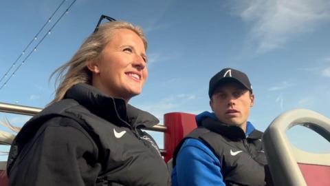 A man and a woman sat on the top deck of an open-top bus. The man is wearing a black cap and looking directly at the camera. The woman is looking into the distance. 