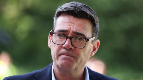 Prime Minister Andy Burnham wears dark-rimmed glasses and a blue suit.