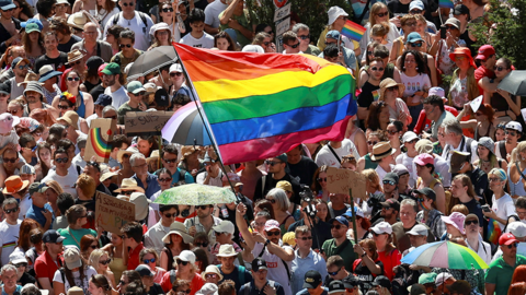 A large group of people stand closely packed, many holding placards and waving rainbow flags—clear signals of LGBTQ+ pride and solidarity.

One prominent sign reads “Je suis” (French for “I am”), while another displays the Hungarian message “A szabadság nem privilégium,” which means “Freedom is not a privilege.”


