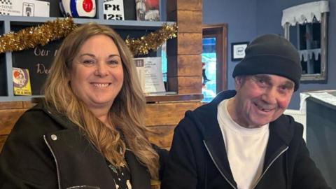 Joanne Percival and John McColl smile as they sit together at a cafe. They wear dark jackets over their tops and there are Mod memorabilia on a display behind