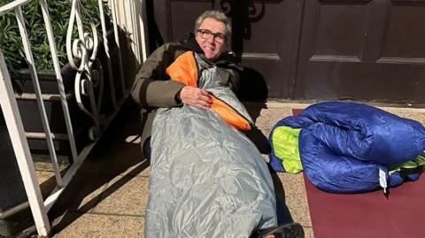 Tim Ringsdore in a grey sleeping bag with orange lining. He is lay on the floor outside a doorway. He is also wearing a khaki coloured coat. He has grey hair and also wears dark square glasses.