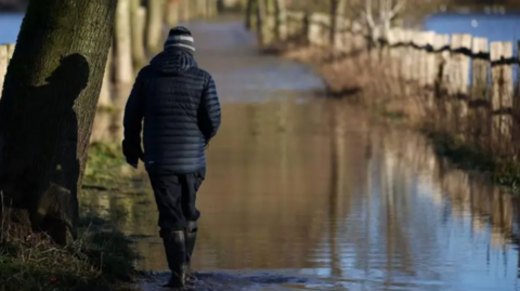 A man walks away from the camera wearing wellies as he walks along a flooden path.