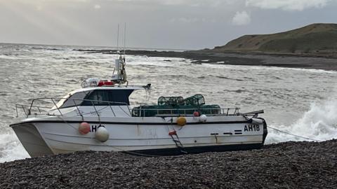 A small white fishing boat with orange buoy attached to it, washed up on a pebbly beach with the waves crashing on to it.