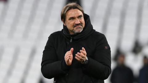 Daniel Farke, wearing a black coat and smiling, applauds at the end of Leeds' FA Cup quarter-final