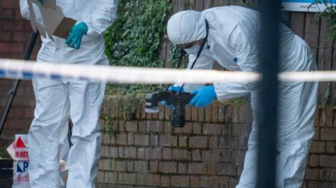 Men in white forensics suits inspect crime scene. One is bent over at the hips taking a photo of evidence.