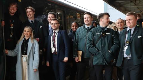 A group of about 10 young people, some dressed in green uniform, next to a train in a station.