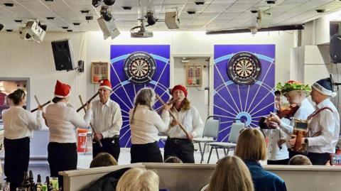 A group of five Morris dancers in white shirts, black trousers and red Christmas hats dancing with sticks in front of an audience. Three other members of the band are playing music. There are dartboards on the wall behind them.