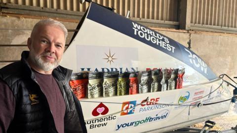 Ollie Phillips smiling at the camera is wearing a burgundy jumper and black gilet with a rowing boat in the background.