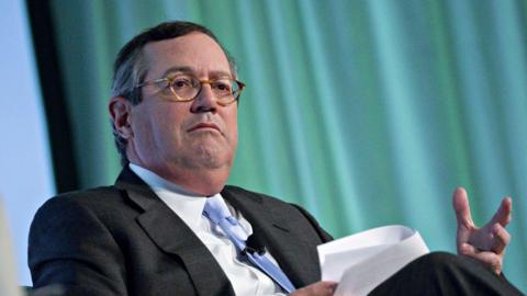 Warren Stephens gestures with his hand as he sits for an interview. He is holding papers in his other hand and wears a black suit, white shirt and light blue tie.