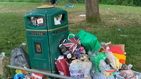 A close up photo of a green bin with the word litter on the site, which has several plastic bags bursting with rubbish propped against it. It is in a grassy location and someone's legs are seen walking behind.