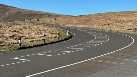 The Mountain Road at Windy Corner. The road rounds a bend an snakes of into the distance, with hills rising on either side and blue sky above.