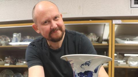 Professor of ceramics Neil Brownsword, holding a blue and white vase, is wearing a black t-shirt and is stood in front of a glass cabinet with a number of ceramic items in it.