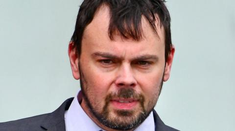 A man wearing a grey suit, purple shirt and tie with white diamonds on it. His top button is undone. He has black hair and hazel eyes and a dark goatie style beard. The background behind him is blue and he is looking down. 