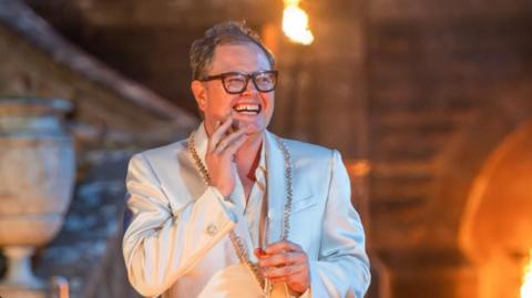 Alan Carr in a white jacket with a jewelled collar and white shirt. He is holding a glass of champagne and his other hand is up to his face. He is smiling and looking to the side of the camera. It is night-time and behind him blurred out is The Celebrity Traitors' castle, with flamed lanterns lighting up the surroundings. Alan is wearing his trademark large framed black glasses and he has short dark hair.