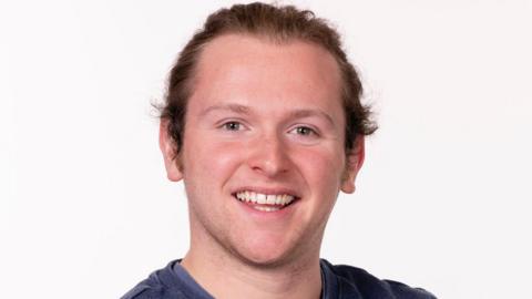 A professional headshot of a man with long brown hair tied back, blond eyebrows, smiling at the camera. He is wearing a bluey grey sweatshirt.