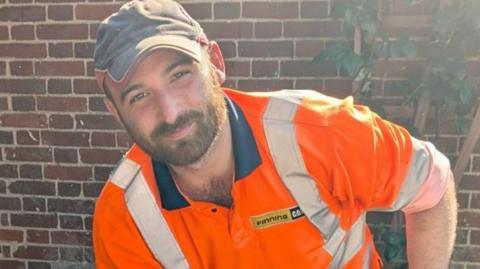 Kieran Palmer standing outside in front of a brick wall. He is looking directly at the camera and smiling and is wearing an orange T-shirt with fluorescent stripes on and is also wearing a baseball cap.