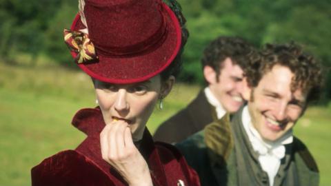 An actress dressed in Georgian clothes eating a primrose covertly, facing away from two men who are laughing behind her