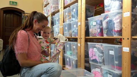 A woman in a pink top and jeans holds her baby daughter in her arm while looking at clothes.