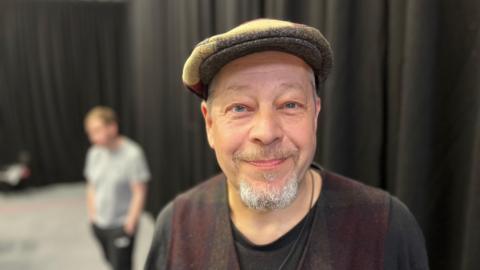 A man with a grey goatee and white hair, he is wearing a flat cap and a vest with a black t-shirt underneath