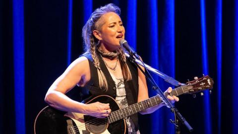KT Tunstall performing on stage in front of a microphone and playing the guitar. She is wearing a black waistcoat and has her hair tied into plaits. 