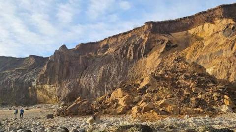 Beige sandstone has collapsed from a cliff at Seaham Hall beach, with two people walking around the landslide.