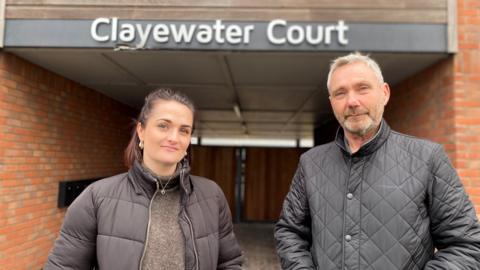 Lucy Tissington and Alan Morris, a young woman and slightly older man, both wearing black padded jackets with their hands in their pockets, standing below a sign that says Clayewater Court