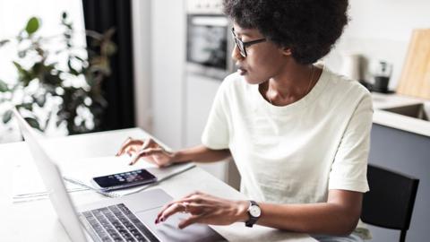 Woman using laptop, notepad, and calculator to look at bills on a white kitchen table