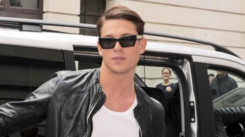 Joey Essex standing outside a taxi, wearing black sunglasses and a black leather jacket. He has brown hair which is swept over.