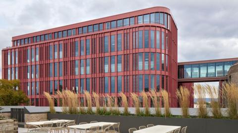 The exterior of the Forum building in Gloucester. The building has a red facade and chairs and tables outside.