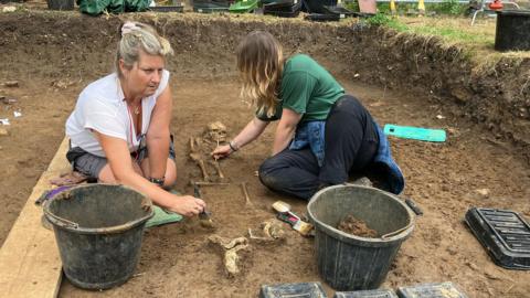 Two women, one with grey hair and wearing a white shirt and grey shorts, and the other with blonde hair and wearing a green top and black trousers, sitting on the floor of an archaeological pit. They're using brushes to brush soil away from a partially-excavated skeleton. They have buckets and tools on the ground around them.